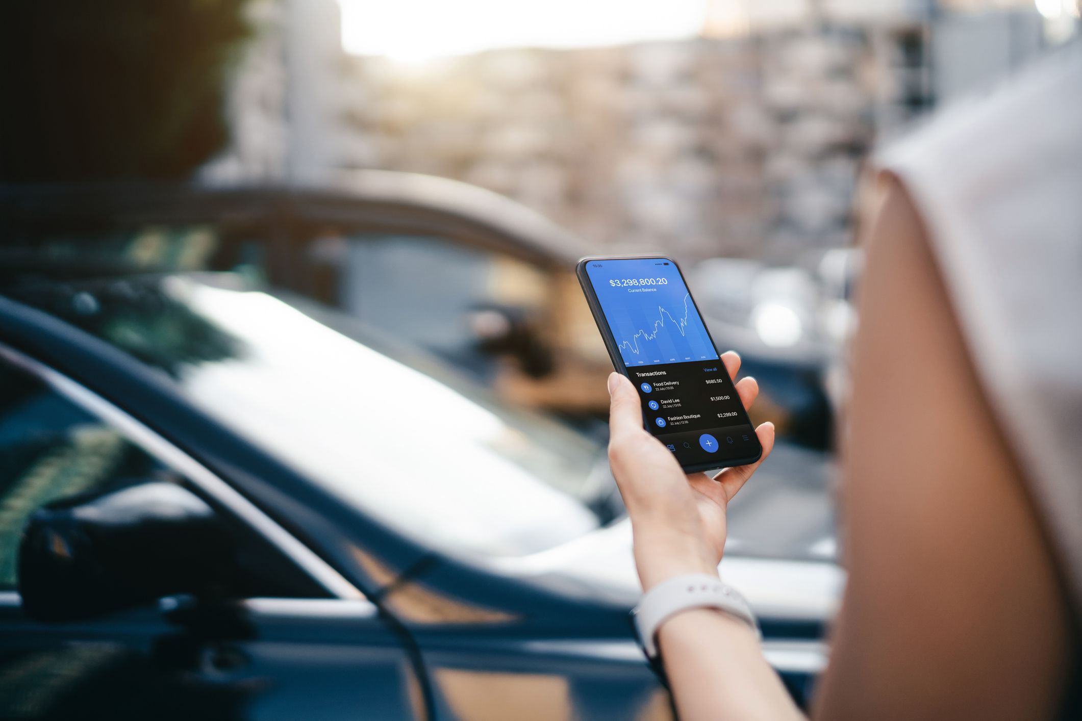 Person hält Smartphone mit geöffneter Finanz-App in der Hand, im Hintergrund ein geparktes Auto.“